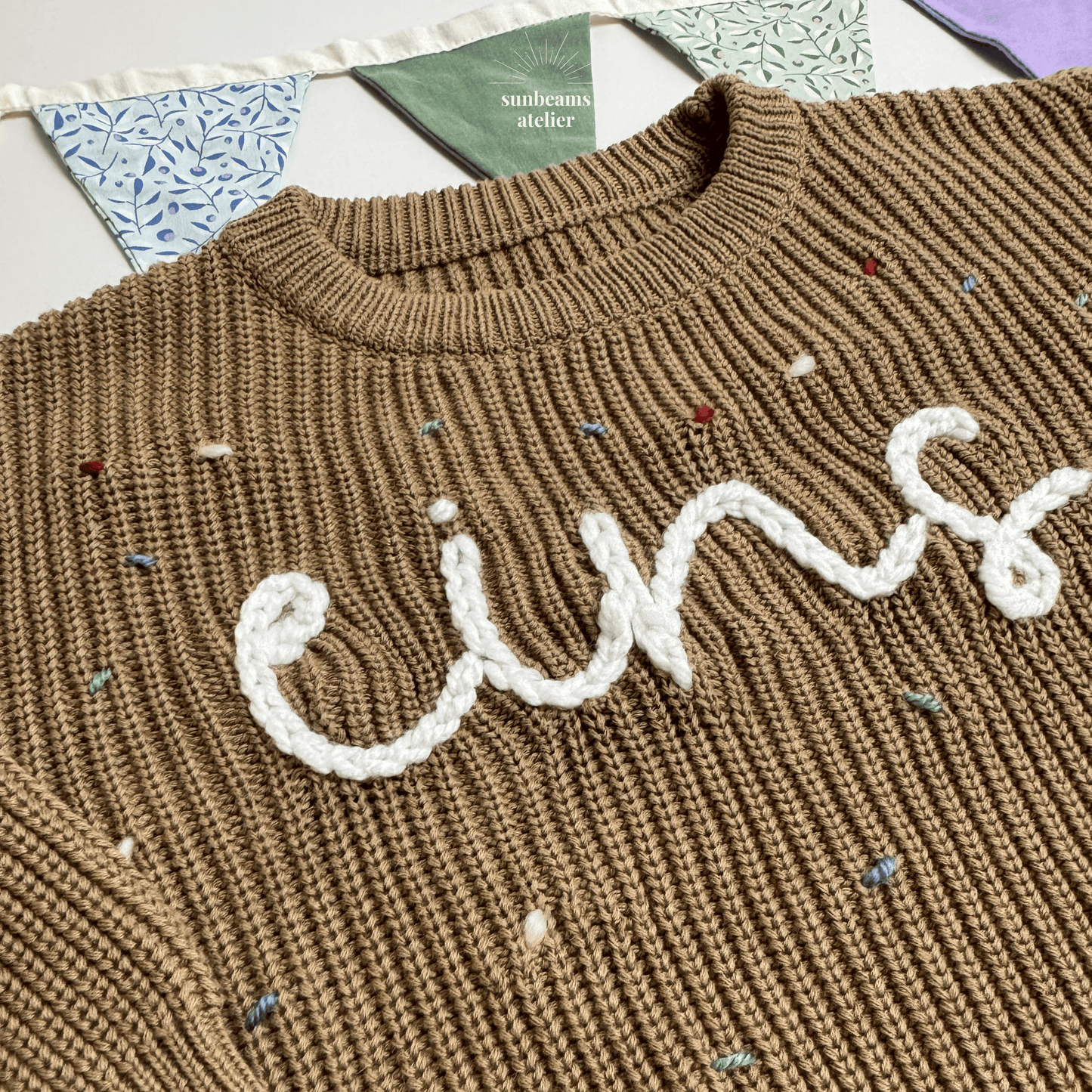 Detailansicht: Tauper Strickpullover mit einem weißen bestickten Schriftzug "eins" zu sehen, außenrum sind bestickte Konfettis