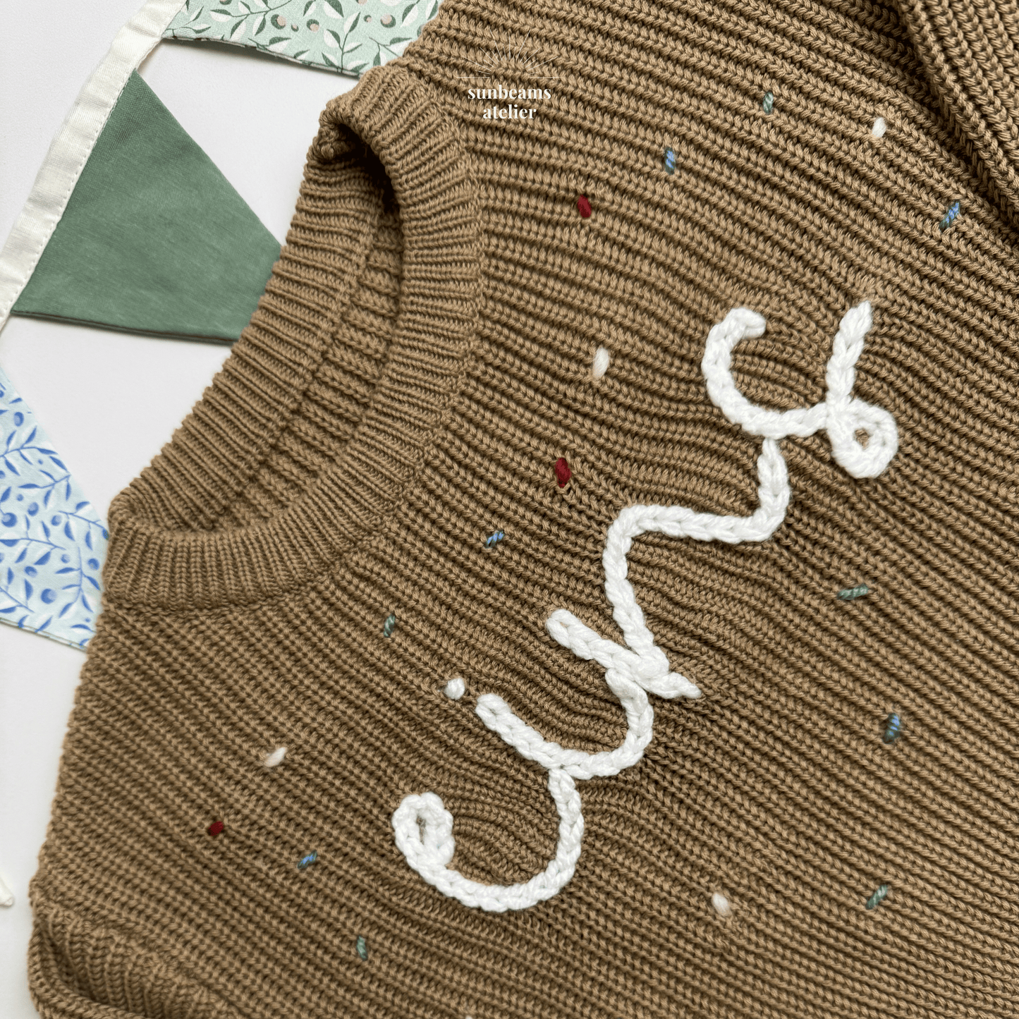 Detailansicht: Tauper Strickpullover mit einem weißen bestickten Schriftzug "eins" zu sehen, außenrum sind bestickte Konfettis