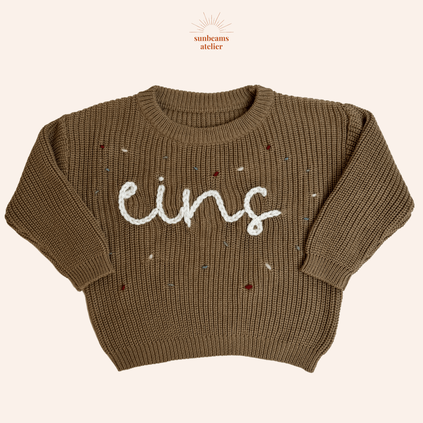 Tauper Strickpullover mit einem weißen bestickten Schriftzug "eins" zu sehen, außenrum sind bestickte Konfettis