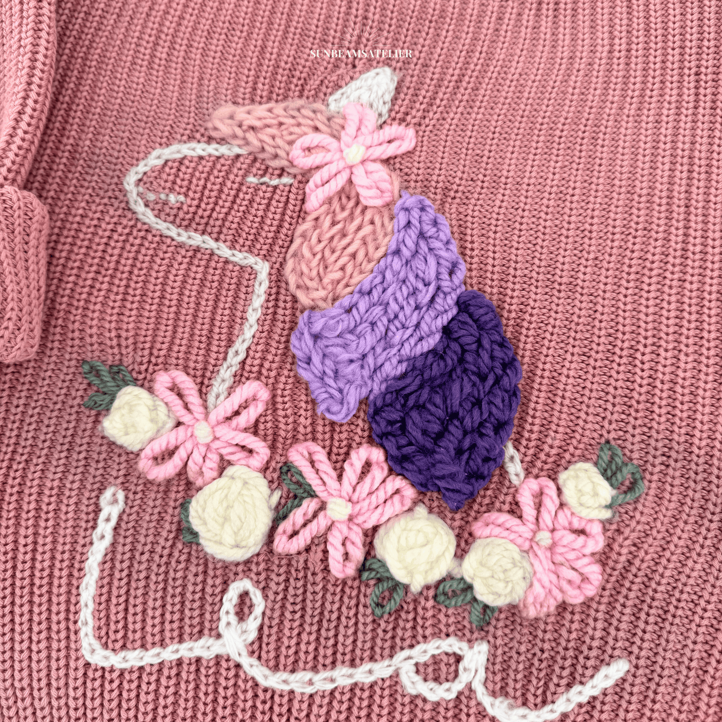 Detailbild: Rosa Strickpullover mit einem handbestickten Einhorn. Die Mähne hat rosa, lavendel und lila Strähnen, oben ist eine bestickte Blume zu sehen sowie ein helles Horn. Das Einhorn hat unten einen bestickten Blumenkranz aus rosa und weißen Blüten/Rosen. Unten ist ein bestickter Schriftzug "Lea" zu sehen