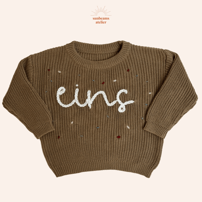 Tauper Strickpullover mit einem weißen bestickten Schriftzug "eins" zu sehen, außenrum sind bestickte Konfettis