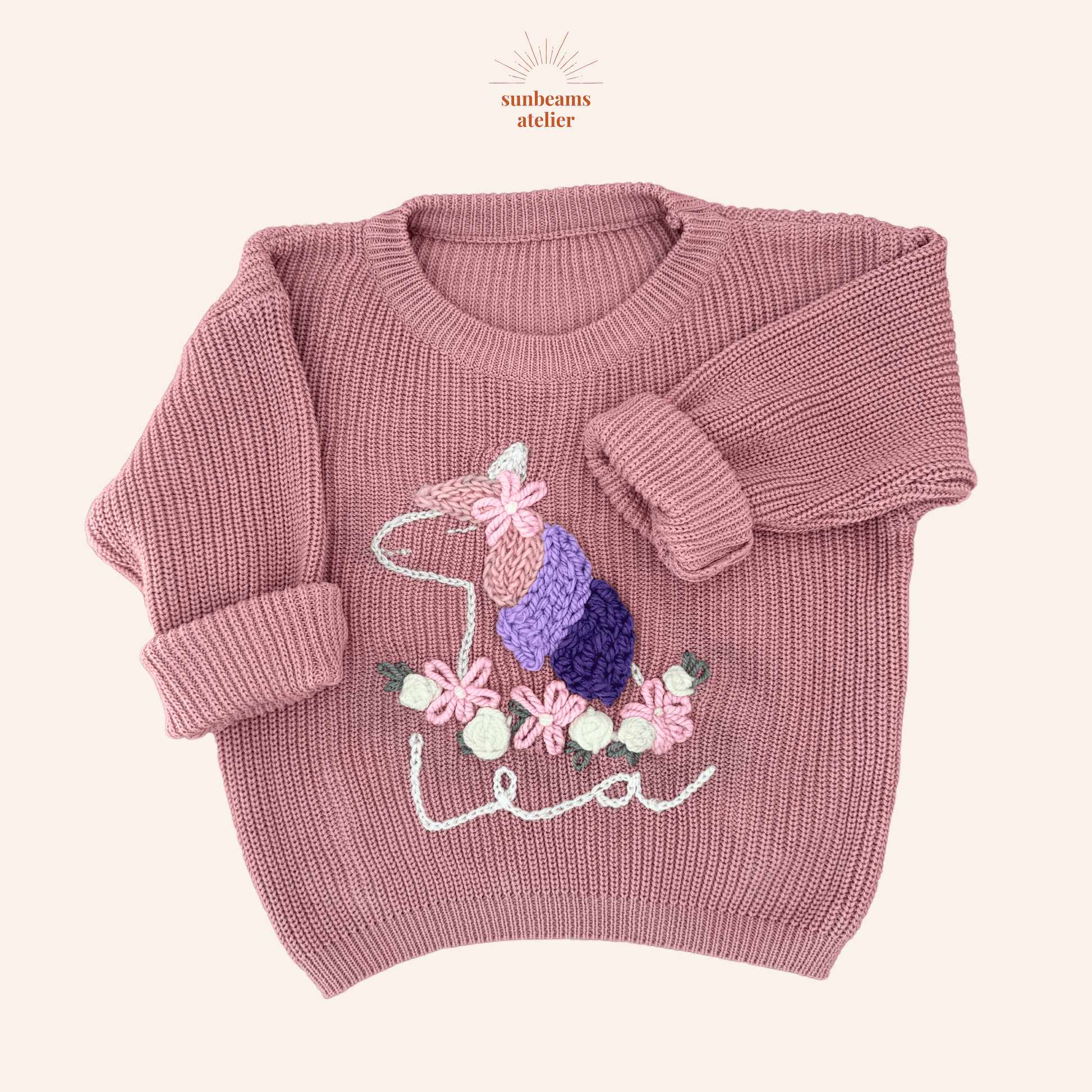Rosa Strickpullover mit einem handbestickten Einhorn. Die Mähne hat rosa, lavendel und lila Strähnen, oben ist eine bestickte Blume zu sehen sowie ein helles Horn. Das Einhorn hat unten einen bestickten Blumenkranz aus rosa und weißen Blüten/Rosen. Unten ist ein bestickter Schriftzug "Lea" zu sehen