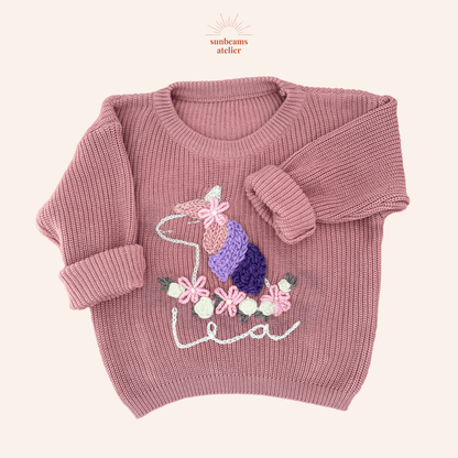 Rosa Strickpullover mit einem handbestickten Einhorn. Die Mähne hat rosa, lavendel und lila Strähnen, oben ist eine bestickte Blume zu sehen sowie ein helles Horn. Das Einhorn hat unten einen bestickten Blumenkranz aus rosa und weißen Blüten/Rosen. Unten ist ein bestickter Schriftzug "Lea" zu sehen
